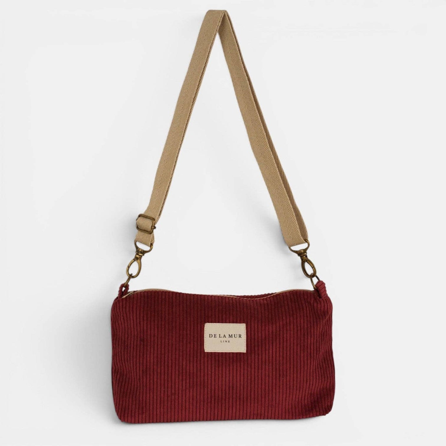 Bordeauxrote De La Mur Arde Clutch aus breitem Cord mit beigefarbenem Tragegurt auf hellem Hintergrund.
