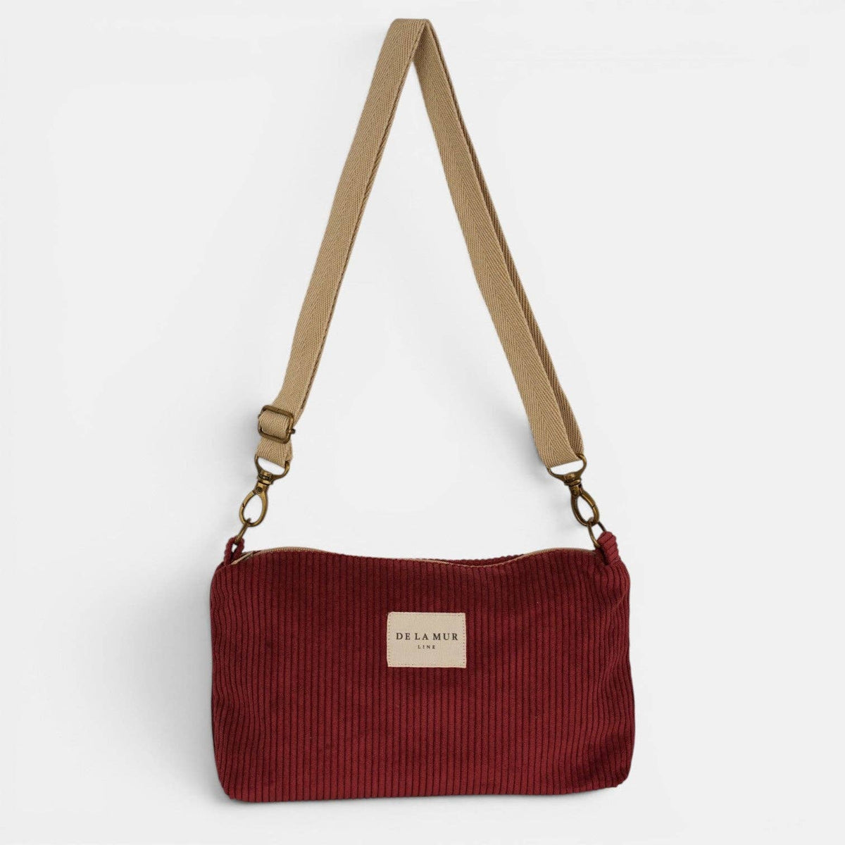 Bordeauxrote De La Mur Arde Clutch aus breitem Cord mit beigefarbenem Tragegurt auf hellem Hintergrund.
