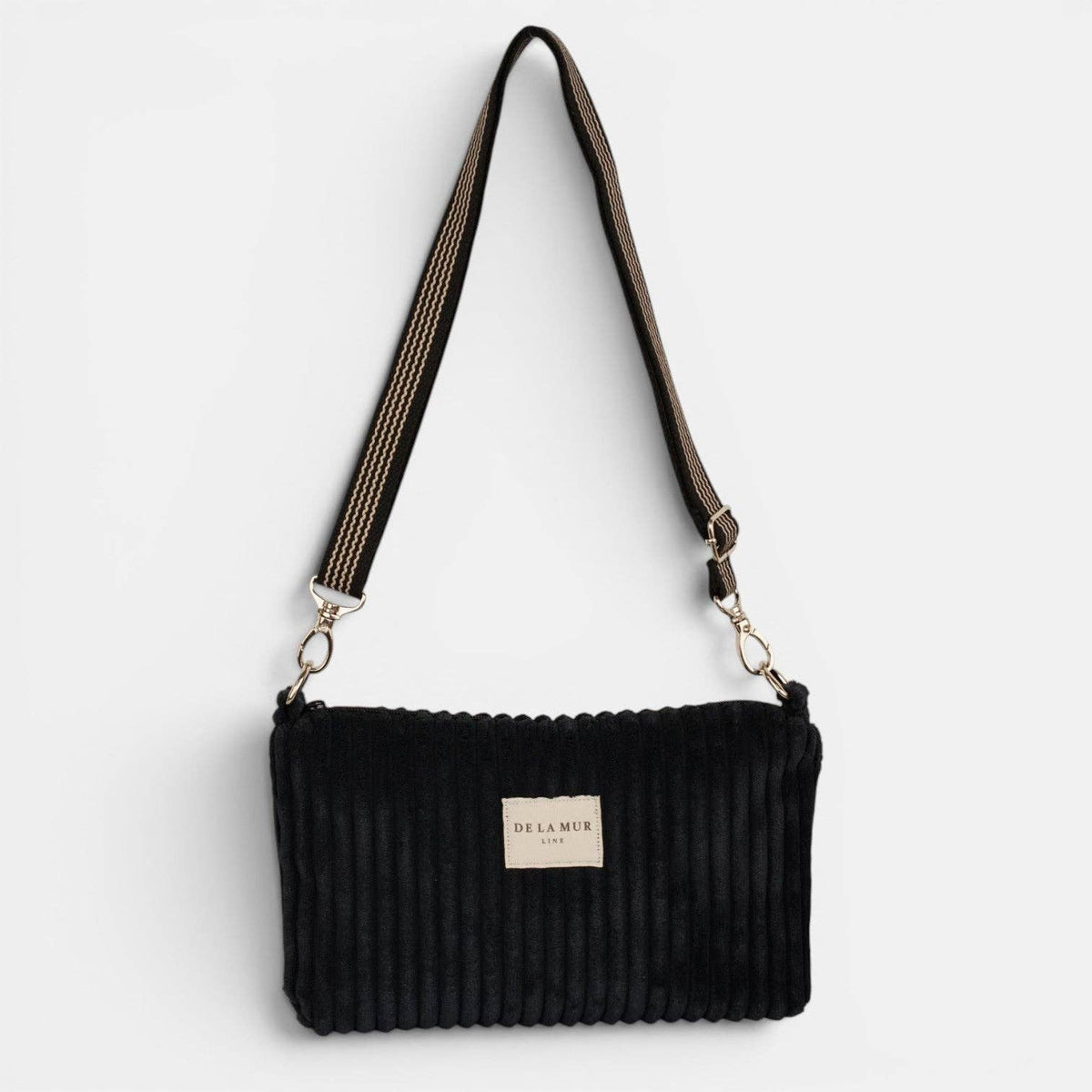 Schwarze De La Mur Curly Clutch aus breitem Cord mit schwarz-beigem Schultergurt auf hellem Hintergrund.