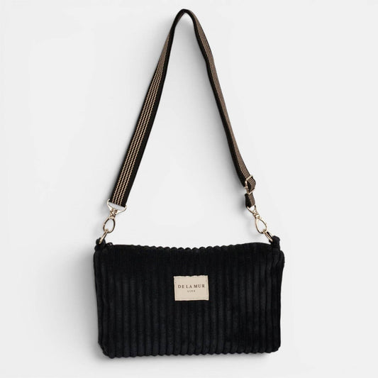 Schwarze De La Mur Curly Clutch aus breitem Cord mit schwarz-beigem Schultergurt auf hellem Hintergrund.